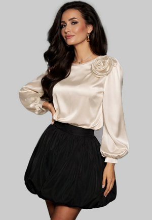 Femme aux longs cheveux foncés portant un chemisier en satin crème avec un détail floral et une jupe bouffante noire, posant avec une main sur la hanche.