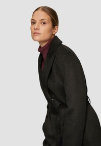 Zwarte wollen jas met een ceintuur in de taille, voorzien van een gelapte revers en zijzakken. Draagt over een bordeauxrode turtleneck.
