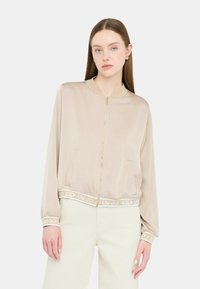Giacca bomber in satin beige con zip frontale, colletto e polsini a costine. Presenta un logo sul fondo e tasche laterali.