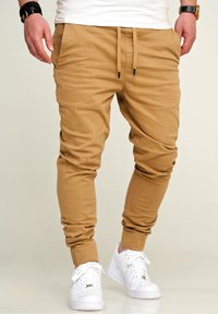 Tansfarbene Jogginghosen aus weichem Stoff, mit elastischen Bündchen und einem Kordelzugbund, kombiniert mit weißen Sneakers.