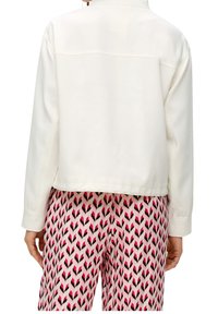 Giacca bianca corta con maniche lunghe e colletto, abbinata a pantaloni rosa con motivi a freccia neri e rossi.