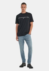 T-shirt nera a maniche corte con la scritta "GAS MILANO 1984" stampata in bianco, abbinata a jeans azzurri chiari e stivaletti neri alla caviglia.