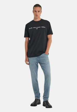T-shirt nera a maniche corte con la scritta "GAS MILANO 1984" stampata in bianco, abbinata a jeans azzurri chiari e stivaletti neri alla caviglia.