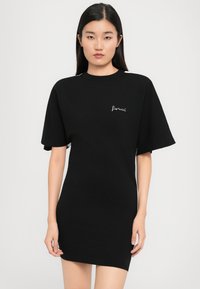 Νεαρή γυναίκα με μακρυά σκούρα μαλλιά, φορώντας έν�α μαύρο στενό κοντομάνικο φόρεμα με το "fiorucci" κεντημένο στο στήθος, στέκεται μπροστά σε λευκό φόντο.