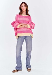 Różowo-żółty sweter z prążkowanym wzorem, z opadającymi ramionami, szerokimi rękawami i luźnym krojem, zestawiony z jasnymi dżinsami i butami w panterkę.