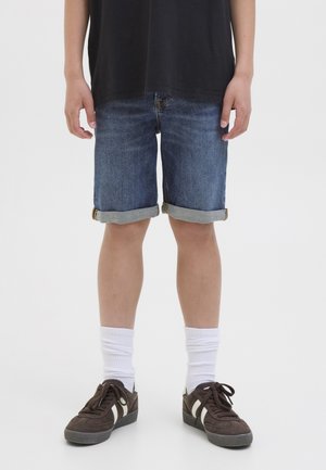 Personne portant un short en jean bleu avec revers, des chaussettes blanches, des baskets marron foncé avec des bandes blanches, et un t-shirt noir, debout sur un fond blanc.