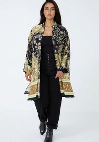 Zwarte en gouden kimono-stijl jas met groene en bruine accenten, voorzien van brede mouwen en een knie-lange zoom. Gecombineerd met zwarte broek en laarzen.
