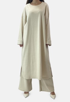 Femme portant une longue tunique côtelée crème avec des fentes latérales, sur un pantalon large assorti et des chaussures à bout pointu, sur un fond uni.