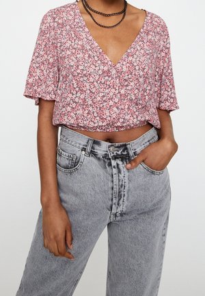 Kobieta ubrana w różowy kwiatowy crop top z głębokim dekoltem w serek oraz jasnoszare wysokie spodnie jeansowe, jedną ręką w kieszeni, nosząca czarny łańcuszkowy naszyjnik.