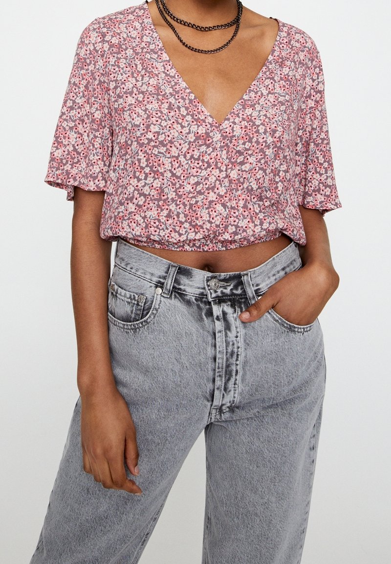Femme portant un crop top fleuri rose avec un col en V profond et un jean taille haute gris clair, une main dans la poche, portant un collier chaîne noir.