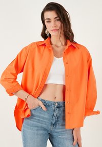 Felle oranje blouse met kraag, lange mouwen opgerold en een losse pasvorm, over een witte cropped tanktop.