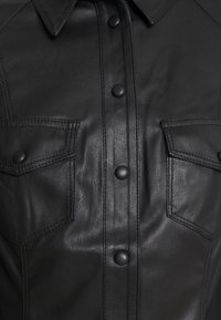 Veste en cuir noire à l'avant avec boutons-pression, deux poches à rabat sur la poitrine avec fermetures à bouton-pression et un col pointu.