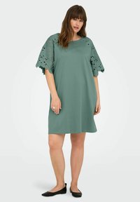 Rochie verde cu o croială lejeră, cu detalii din dantelă la mânecile scurte. Material neted și design până la genunchi. Sandale negre completează look-ul.