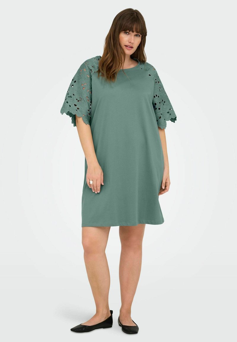 Rochie verde cu o croială lejeră, cu detalii din dantelă la mânecile scurte. Material neted și design până la genunchi. Sandale negre completează look-ul.