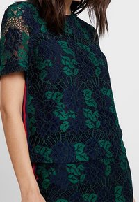 Spetsblus med blommönster i grönt och marinblått, med korta ärmar och röda accenter längs sidorna. Texturerad och lager-på-lager design.