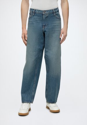 Personne portant un jean denim bleu ample et des baskets blanches, debout devant un fond uni.