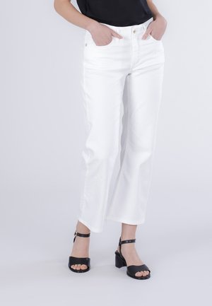 Persona che indossa jeans bianchi a gamba dritta e sandali neri aperti con tacco, mani in tasca, su uno sfondo chiaro uniforme.