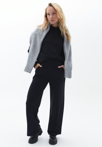 Grå stickad cardigan över en svart långärmad topp, parat med svarta vida byxor och svarta skor. Släta texturer och enkel design.