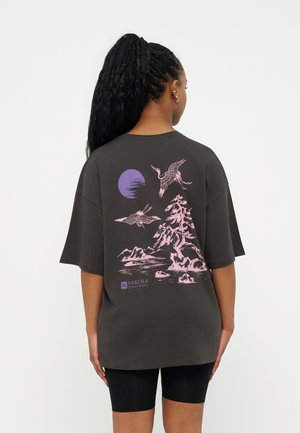 Γυναίκα με μακριά πλεξούδα φοράει σκούρο, φαρδύ μπλουζάκι που δείχνει ροζ γερανούς που πετούν πάνω από ροζ δέντρα και βουνά κάτω από μοβ ήλιο.