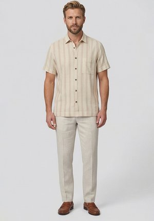 Man draagt een beige overhemd met korte mouwen en strepen, lichtbeige broek en bruine leren loafers, staand tegen een effen achtergrond.