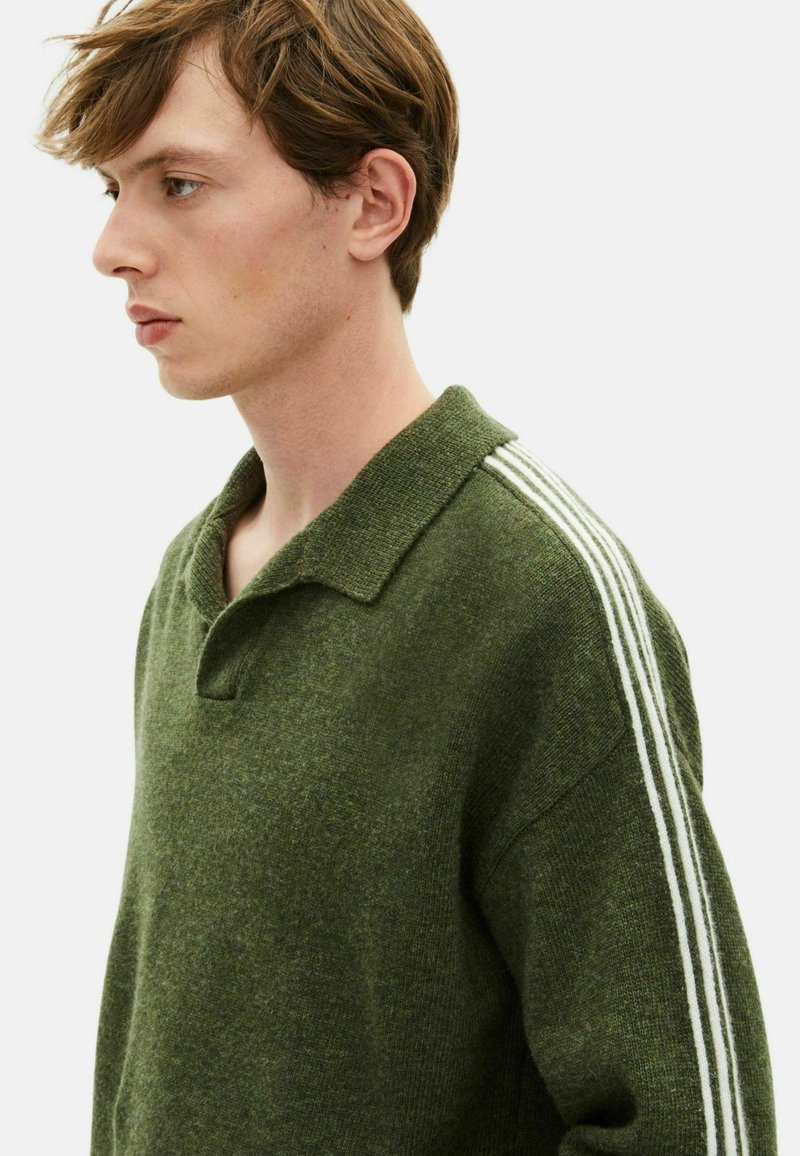 Jeune homme en pull en tricot vert avec des rayures blanches sur les épaules, regardant à gauche, sur fond uni clair.