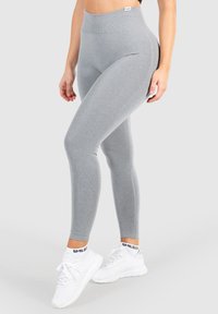 Šedé legíny vyrobené z pružného materiálu s vysokým pasem, hladkou texturou a přiléhavým střihem. Nosené s bílými sportovními botami.