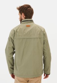 Olive green jacket with a relaxed fit, buttoned collar, and a brown leather logo patch on the upper back. Smooth, lightweight fabric.