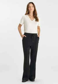 Jonge vrouw met lang bruin haar, gekleed in een witte T-shirt met korte mouwen en een V-hals, en zwarte broek met trekkoorden, staande met handen in haar zakken.
