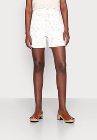 Persona que lleva pantalones cortos blancos de cintura alta con estampado floral y sandalias marrones de tacón abiertas, de pie contra un fondo liso y claro.