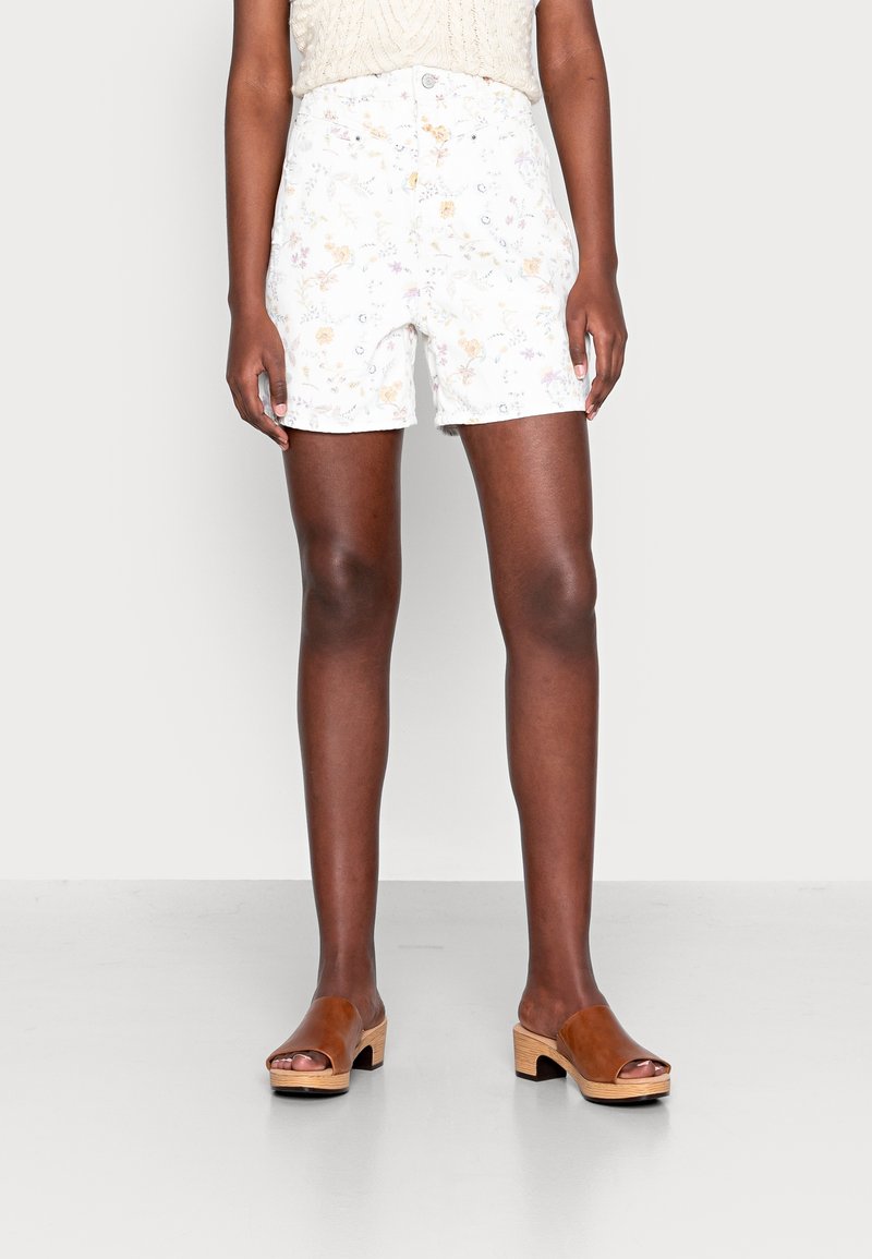 Persona que lleva pantalones cortos blancos de cintura alta con estampado floral y sandalias marrones de tacón abiertas, de pie contra un fondo liso y claro.