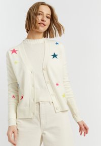 Cardigan di crema chiara con motivi a stelle colorate in rosa, verde, blu e blu scuro. Presenta bottoni e polsini a coste, indossato sopra un maglione.