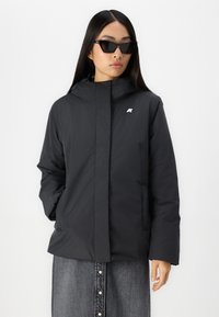 Giacca impermeabile nera con cappuccio, chiusura frontale con zip, tasche laterali e un piccolo logo sul petto; texture liscia e design minimale.