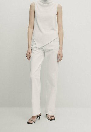 Femme portant un haut asymétrique blanc sans manches, un pantalon droit blanc et des sandales noires à talons et à brides, debout.