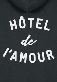 Tmavě šedá mikina s kapucí, na které je bílé vyšívané text: "HÔTEL de l'amour." Text je umístěn ve středové zakřivené kompozici. Měkká textura.