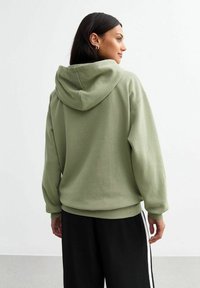 Sudadera con capucha de gran tamaño en algodón verde claro, con un bolsillo canguro, puños y dobladillo de canalé. Combinada con pantalones negros con rayas laterales.