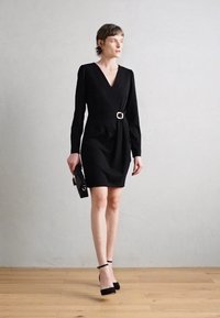 Robe noire portefeuille à manches longues, décolleté en V et détail de ceinture. Le modèle tient une pochette noire et porte des chaussures à talons hauts noires. Texture lisse.