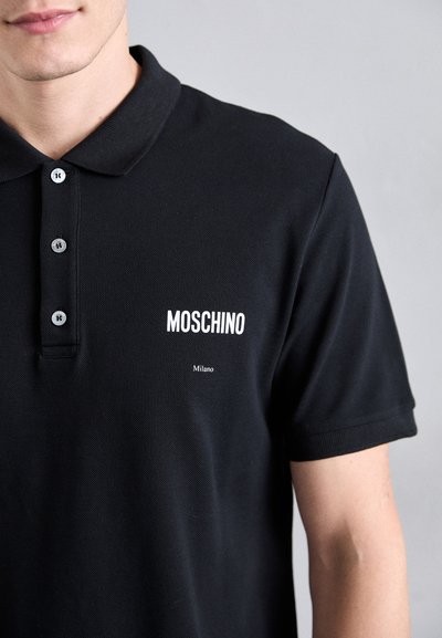 Juoda polo marškinėliai, pagaminti iš tekstūruotos medžiagos, turi tris mygtukus ir baltą „MOSCHINO“ bei „Milano“ tekstą kairiajame krūtinės kampe.
