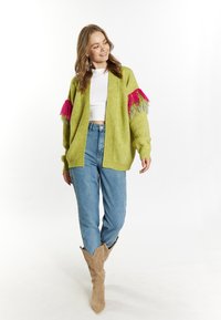 Lindgrünem Cardigan mit rosa und grauen Fransenakzenten an den Ärmeln, getragen mit hellblauer Jeans und beigefarbenen Cowboystiefeln.