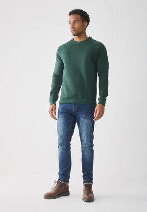 Man som står, klädd i en grön sweatshirt, blå jeans och bruna snörade kängor, mot en enkel ljus bakgrund.