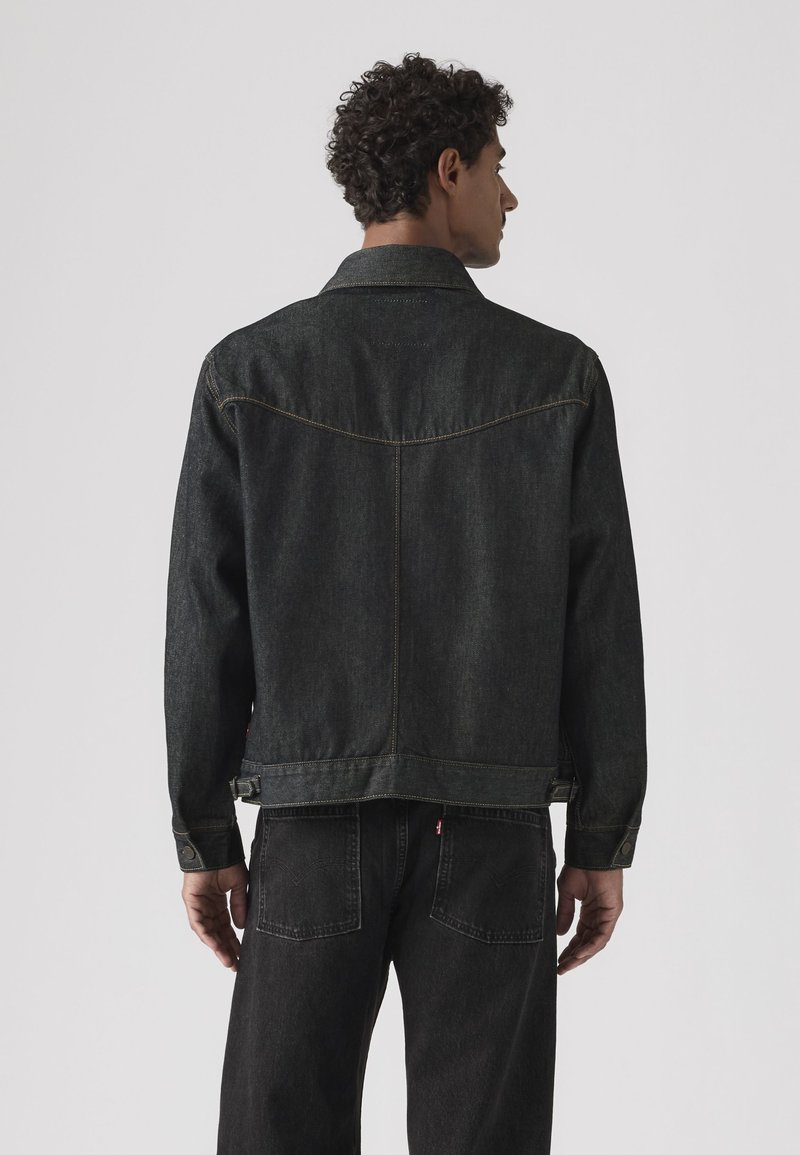 Mand med krøllet hår iført en sort denimjakke og sorte jeans, står med ryggen mod en ensfarvet lys baggrund.