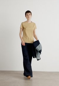 Tans kleurig katoenen t-shirt met een ronde halslijn, gecombineerd met donkerblauwe flared jeans en een opgevouwen spijkerjacket, vastgehouden in de linkerarm.