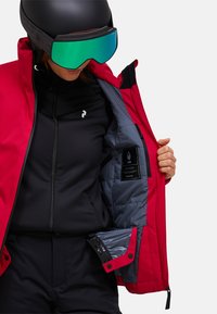 Persoon in een zwarte skikleding, met een open rode jas die de gevoerde grijze binnenkant laat zien, een zwarte helm en reflecterende groene skibril.