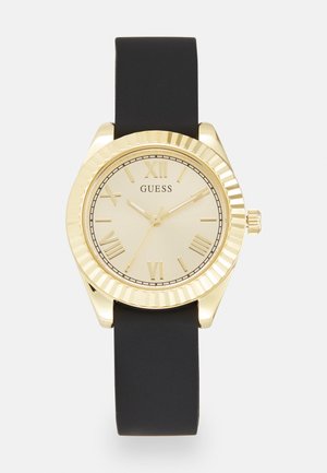 Montre dorée avec un cadran rond, un bracelet en caoutchouc noir, des chiffres romains et une lunette texturée. Présente un design minimaliste avec un cadran simple.