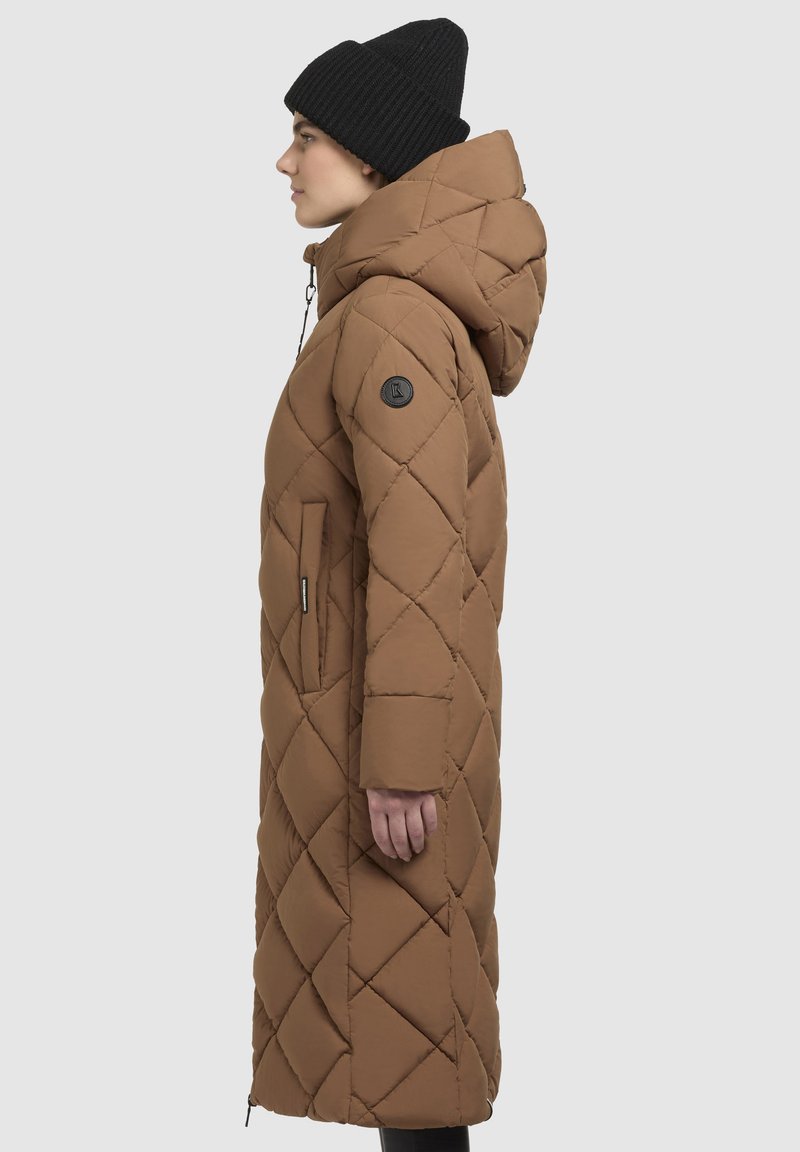 Langer brauner gesteppter Daunenmantel mit Kapuze, Reißverschlusstaschen und einem strukturierten Stoff mit Rautenmuster. Schwarze Strickmütze auf dem Kopf.