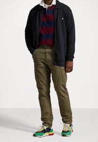 Man som bär en marinblå zip-up jacka, en burgunder och marinrandig polo-tröja, olivgröna cargo-byxor, flerfärgade sneakers och ett grönrandigt bälte.