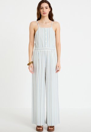 Kvinde iført en ærmeløs, knappet, stribet jumpsuit med vidde bukseben og brune platformsandaler, stående mod en hvid baggrund.