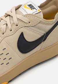 Beige Nike-sneakers med svart swoosh, gula detaljer, beige snörningar, och en strukturad överdel i mocka och nät.