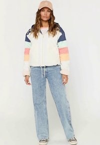 Chaqueta de forro polar colorida con cierre frontal de cremallera, que presenta cuerpo blanco, mangas multicolor a rayas y una textura acogedora. Combinada con jeans azul claro.