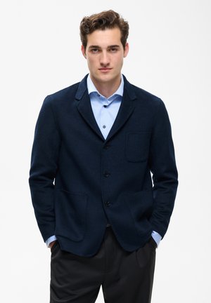 Jeune homme aux cheveux bruns courts portant un blazer bleu marine foncé sur une chemise bleu clair, debout les mains dans les poches devant un fond blanc.