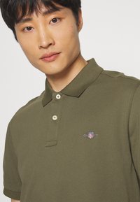Olivengrøn polo-shirt med krave, to-knaps placket og broderet logo på brystet. Fremstillet af struktureret bomuldsstof.